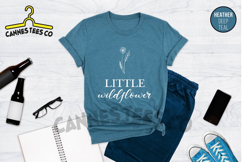 K&ouml;nnte beinhalten: Ein blaues T-Shirt mit einem wei&szlig;en Umriss einer Blume und dem Text "LITTLE wildflower".