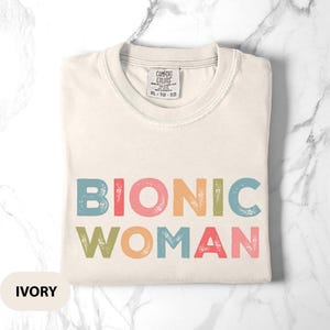 May include: Ivory-colored t-shirt with the words "BIONIC WOMAN" in colorful, retro-style lettering. The shirt has a crew neck and a tag that reads "COMFORT COLORS". The shirt is folded neatly on a marble surface.