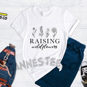 K&ouml;nnte beinhalten: Wei&szlig;es T-Shirt mit einer schwarzen Grafik von Blumen und dem Text "Raising wildflowers".