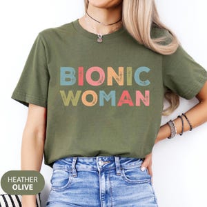 May include: A green t-shirt with the text "Bionic Woman" in colorful, distressed letters. The shirt is made of a soft, comfortable material and has a relaxed fit.