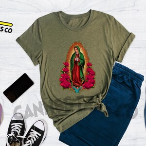 May include: A heather olive t-shirt featuring a graphic of Our Lady of Guadalupe surrounded by red roses. The t-shirt is shown on a model with blue jeans and a backpack.