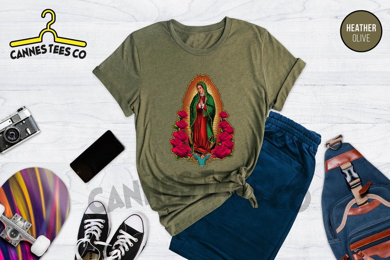 May include: A heather olive t-shirt featuring a graphic of Our Lady of Guadalupe surrounded by red roses. The t-shirt is shown on a model with blue jeans and a backpack.