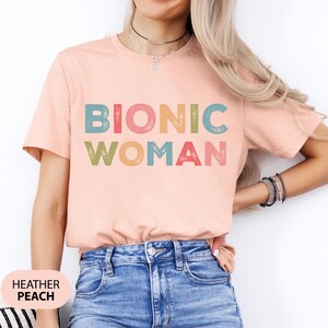 May include: A heather peach t-shirt with the text "Bionic Woman" in colorful, distressed letters.
