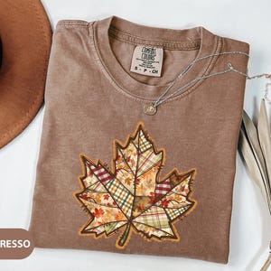May include: Espresso-colored t-shirt with a large, patchwork-style leaf design. The leaf features various plaid and floral patterns in fall colors. A silver necklace with a small pendant rests on the shirt. The shirt is labeled "Comfort Colors".