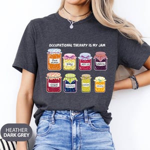 May include: A heather dark grey t-shirt with the text "Occupational Therapy is My Jam" and a graphic of eight jars of jam, each representing a different aspect of occupational therapy, such as fine motor, play, ADLs, learn, visual motor, cognition, social, and sensory.