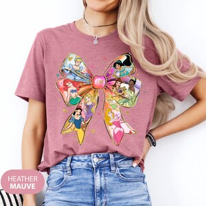 May include: A heather mauve t-shirt featuring a large bow with various Disney princesses inside. The princesses include Cinderella, Ariel, Belle, Snow White, Aurora, and more.