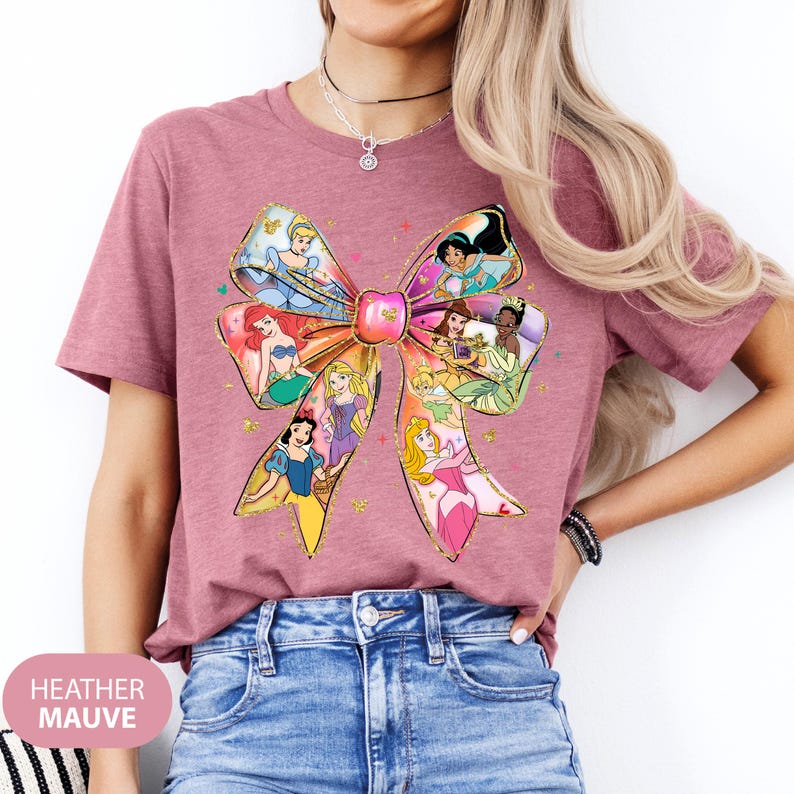 May include: A heather mauve t-shirt featuring a large bow with various Disney princesses inside. The princesses include Cinderella, Ariel, Belle, Snow White, Aurora, and more.