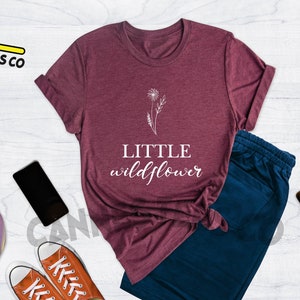 K&ouml;nnte beinhalten: T-Shirt in Heide-Maroon mit einem wei&szlig;en L&ouml;wenzahnmuster und dem Text "LITTLE wildflower".