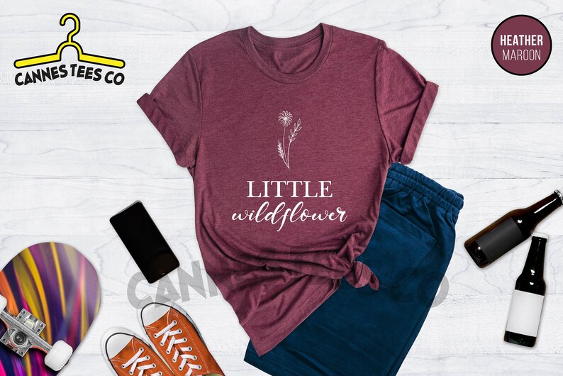 K&ouml;nnte beinhalten: T-Shirt in Heide-Maroon mit einem wei&szlig;en L&ouml;wenzahnmuster und dem Text "LITTLE wildflower".