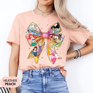 May include: A peach coloured t-shirt with a large bow design featuring various Disney princesses. The bow is made of gold glitter and the princesses are in their iconic dresses. The shirt is made of a soft, comfortable material.