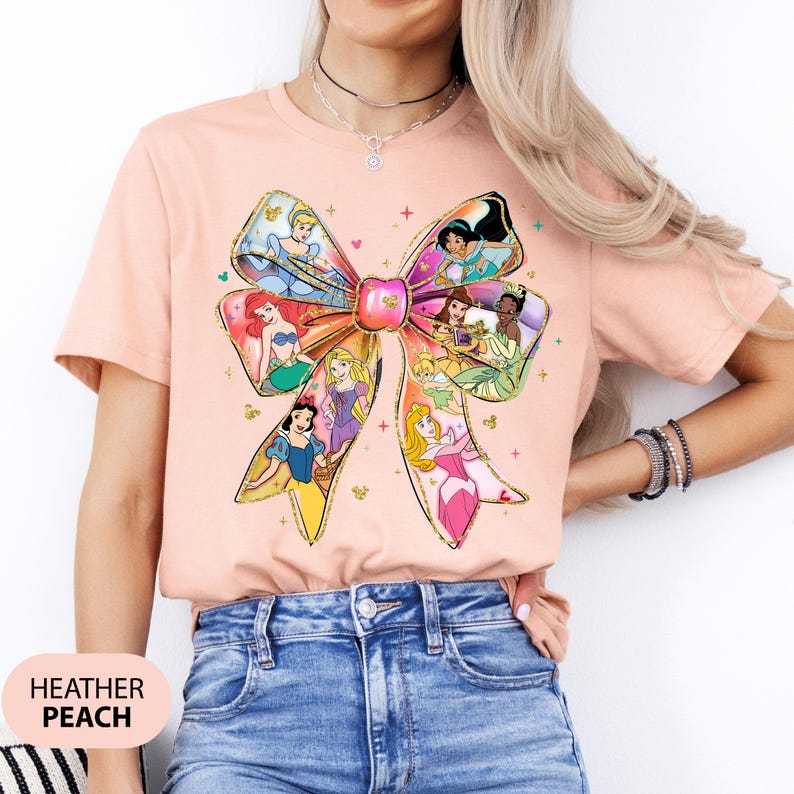 May include: A peach coloured t-shirt with a large bow design featuring various Disney princesses. The bow is made of gold glitter and the princesses are in their iconic dresses. The shirt is made of a soft, comfortable material.