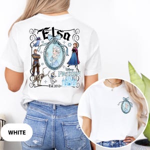 May include: White t-shirt featuring a Disney Frozen design. The back of the shirt displays the word "Elsa" above a framed image of Elsa, with other characters and the text "Frozen" and "Est. 2013". A smaller version of the design is on the front.