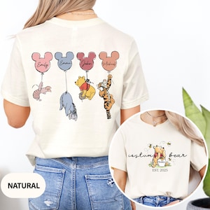 May include: A white t-shirt with a design of Winnie the Pooh and friends holding balloons with names on them. The back of the shirt says "custom bear est. 2025".