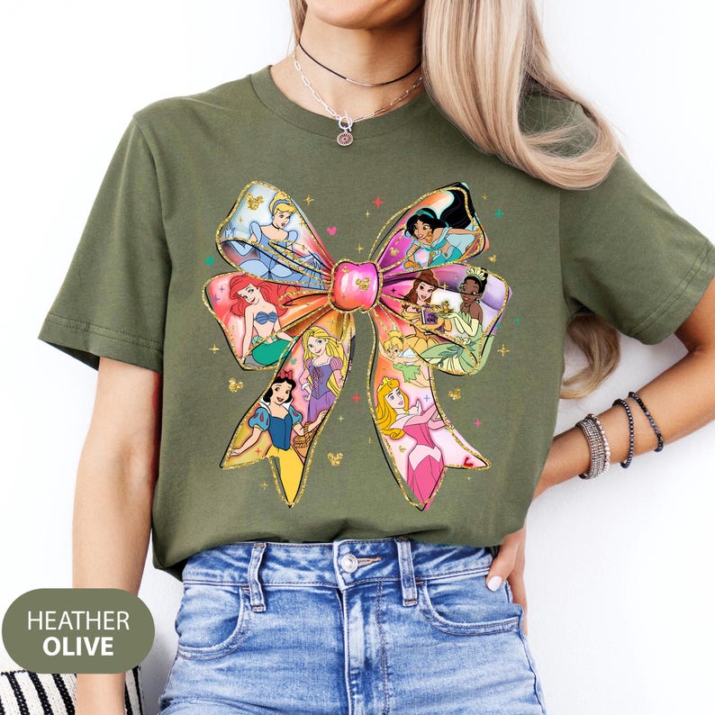 May include: A green t-shirt featuring a large bow design with various Disney princesses. The bow is made up of different coloured ribbons and each princess is depicted in her signature dress. The shirt is made of a soft, comfortable material and is perfect for any Disney fan.