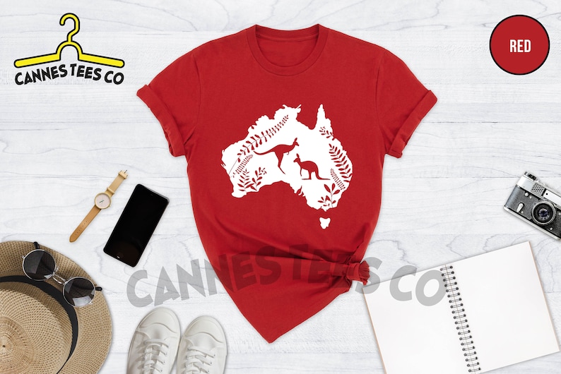 Może przedstawiać: Czerwona koszulka z białą sylwetką kontynentu australijskiego z dwoma kangurami i roślinnością. Na koszulce nadrukowany jest tekst "Cannes Tees Co".