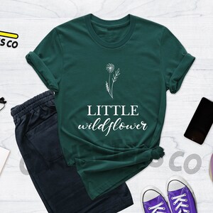 K&ouml;nnte beinhalten: Ein dunkelgr&uuml;nes T-Shirt mit einer wei&szlig;en Umrandung eines L&ouml;wenzahns und dem Text "LITTLE wildflower" in Wei&szlig;.
