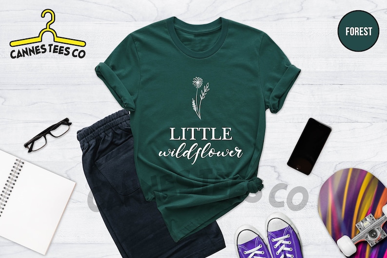 K&ouml;nnte beinhalten: Ein dunkelgr&uuml;nes T-Shirt mit einer wei&szlig;en Umrandung eines L&ouml;wenzahns und dem Text "LITTLE wildflower" in Wei&szlig;.