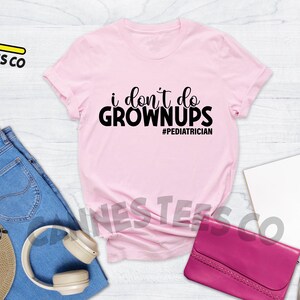 May include: Pink t-shirt with black text that reads "i don't do grown ups #pediatrician". The shirt is laid flat on a white wooden surface with jeans, a hat, headphones, a purse, and sneakers.
