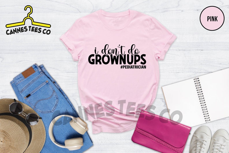 May include: Pink t-shirt with black text that reads "i don't do grown ups #pediatrician". The shirt is laid flat on a white wooden surface with jeans, a hat, headphones, a purse, and sneakers.