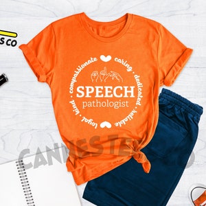 Op de afbeelding: Oranje T-shirt met een wit cirkelvormig ontwerp met de tekst "Speech Pathologist" met handgebaren voor het woord "speech" in de cirkel. De cirkel is omgeven door de woorden "compassionate, caring, dedicated, reliable, loyal, kind".