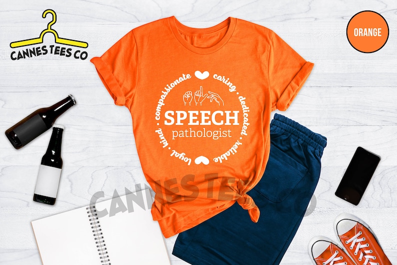Op de afbeelding: Oranje T-shirt met een wit cirkelvormig ontwerp met de tekst "Speech Pathologist" met handgebaren voor het woord "speech" in de cirkel. De cirkel is omgeven door de woorden "compassionate, caring, dedicated, reliable, loyal, kind".