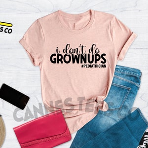 May include: A pink t-shirt with the text "i don't do grown ups #pediatrician" printed in black. The shirt is laid out on a white wooden surface with a pair of blue jeans, a purse, sunglasses, a cell phone, a watch, and a camera.