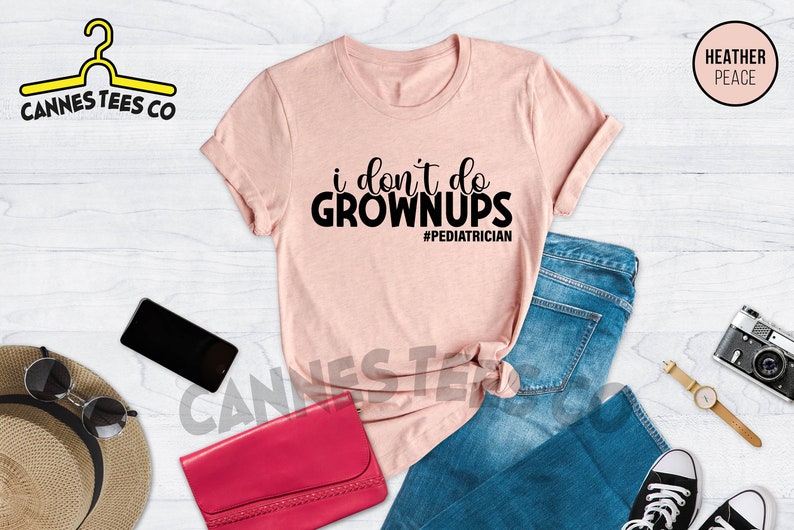 May include: A pink t-shirt with the text "i don't do grown ups #pediatrician" printed in black. The shirt is laid out on a white wooden surface with a pair of blue jeans, a purse, sunglasses, a cell phone, a watch, and a camera.