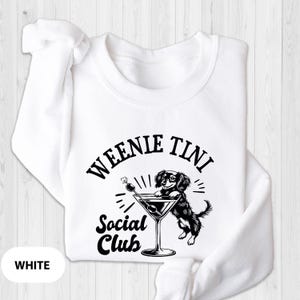 May include: White sweatshirt with a black and white graphic of a dachshund dog holding a martini glass. The text "Weenie Tini Social Club" is printed on the sweatshirt.