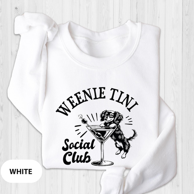May include: White sweatshirt with a black and white graphic of a dachshund dog holding a martini glass. The text "Weenie Tini Social Club" is printed on the sweatshirt.