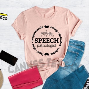 Op de afbeelding: Een roze T-shirt met een zwart cirkelontwerp met de tekst "Speech Pathologist" en een gebarentaal symbool in het midden. De cirkel is omringd door de woorden "compassionate, caring, dedicated, reliable, loyal, kind".