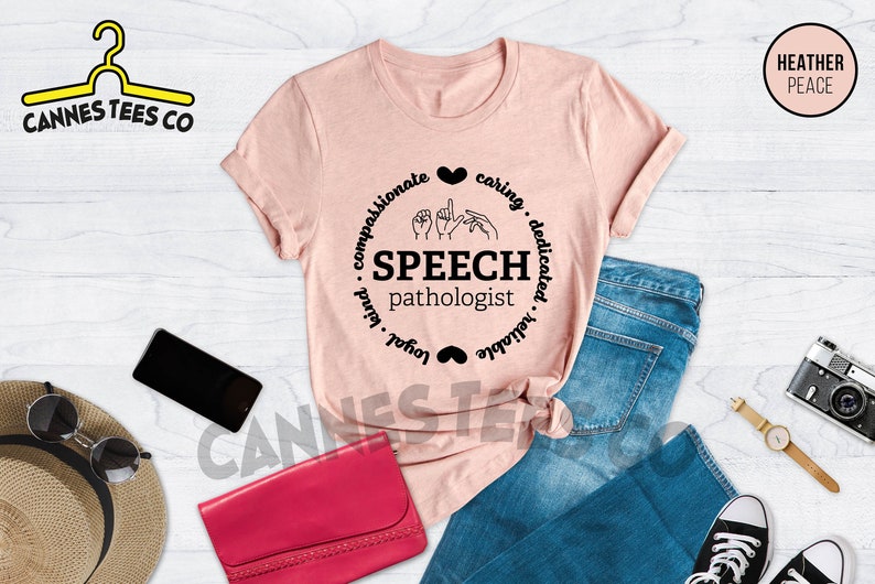 Op de afbeelding: Een roze T-shirt met een zwart cirkelontwerp met de tekst "Speech Pathologist" en een gebarentaal symbool in het midden. De cirkel is omringd door de woorden "compassionate, caring, dedicated, reliable, loyal, kind".