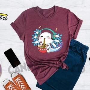 May include: Heather maroon t-shirt featuring a cartoon axolotl wearing headphones and eating ramen noodles. The axolotl is surrounded by blue and green water plants. The text "Cannes Tees Co" is visible in the top left corner of the image.