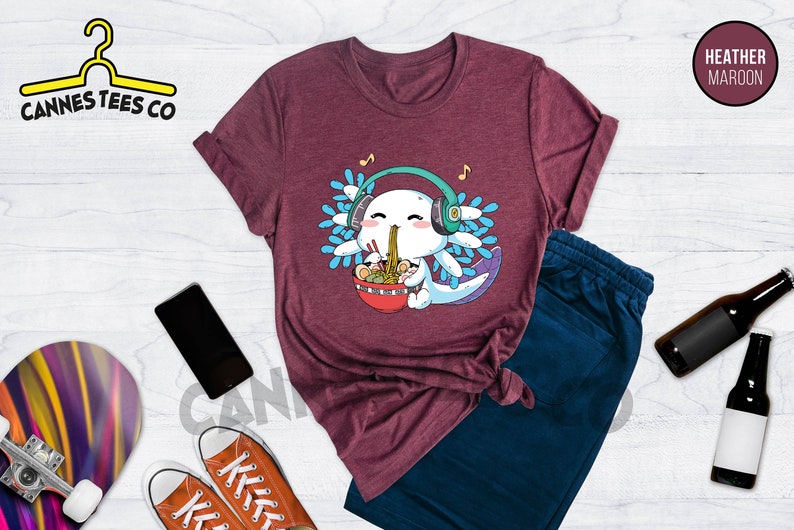 May include: Heather maroon t-shirt featuring a cartoon axolotl wearing headphones and eating ramen noodles. The axolotl is surrounded by blue and green water plants. The text "Cannes Tees Co" is visible in the top left corner of the image.