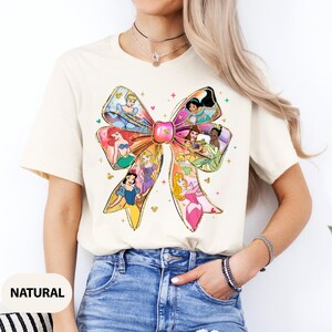 May include: A white t-shirt featuring a colourful bow design with various Disney princesses. The bow is made up of different coloured ribbons with each princess depicted in a different section of the bow. The princesses include Cinderella, Ariel, Belle, Snow White, Aurora, and Tiana.