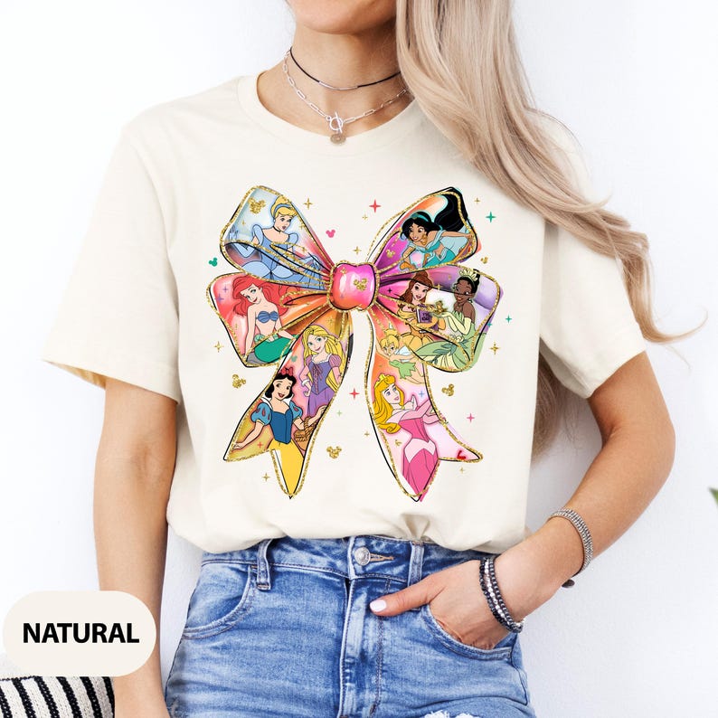 May include: A white t-shirt featuring a colourful bow design with various Disney princesses. The bow is made up of different coloured ribbons with each princess depicted in a different section of the bow. The princesses include Cinderella, Ariel, Belle, Snow White, Aurora, and Tiana.