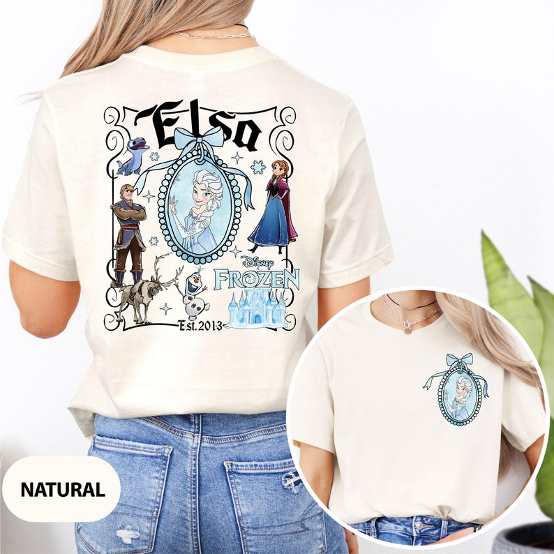May include: Cream-colored t-shirt featuring a Disney Frozen design. The back of the shirt displays the name "Elsa" in a decorative frame with character illustrations. A smaller version of the design is on the front.
