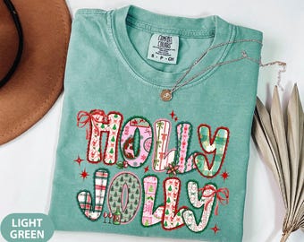 May include: Light green t-shirt with the words "HOLLY JOLLY" in a festive design. The letters are decorated with Christmas patterns, bows, and stars. A silver necklace with a pendant is draped across the shirt.