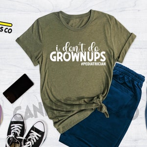 May include: A green t-shirt with the text "i don't do grownups #pediatrician" in white. The t-shirt is on a white wooden surface with a pair of blue sweatpants, a skateboard, a camera, a phone, and a blue backpack.