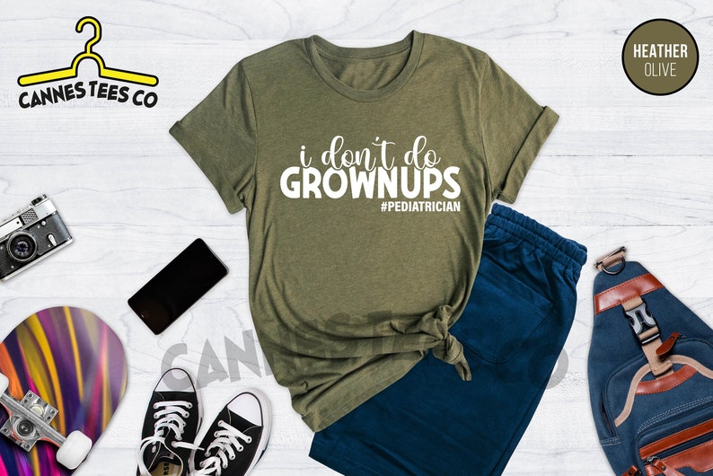 May include: A green t-shirt with the text "i don't do grownups #pediatrician" in white. The t-shirt is on a white wooden surface with a pair of blue sweatpants, a skateboard, a camera, a phone, and a blue backpack.