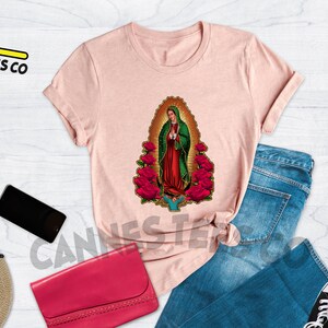May include: A pink t-shirt with a graphic of Our Lady of Guadalupe surrounded by red roses. The text "HEATHER PEACE" is in the upper right corner of the image.