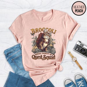 May include: A pink heather t-shirt with a graphic of a cat wearing a backpack and a crown. The cat is standing on a stone staircase with a castle in the background. The text "Broccoli Official Mascot of Quest Squad" is printed below the cat.