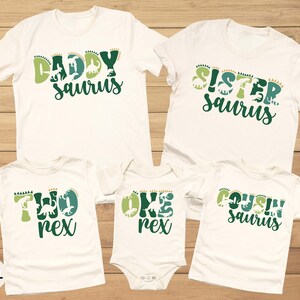 May include: White t-shirts and a onesie with green and light green dinosaur-themed text. The shirts read "Daddy Saurus", "Sister Saurus", "Two Rex", "One Rex", and "Cousin Saurus". The shirts are displayed on a wooden surface.