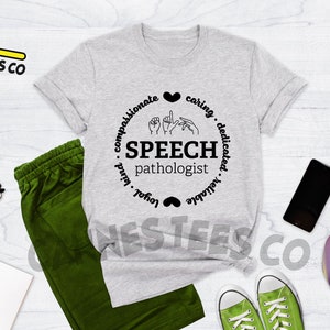 Op de afbeelding: Grijs T-shirt met een zwart cirkelontwerp met de woorden "SPEECH pathologist" en gebarentaal symbolen. De cirkel is omringd door de woorden "compassievol, zorgzaam, toegewijd, betrouwbaar, loyaal, vriendelijk".