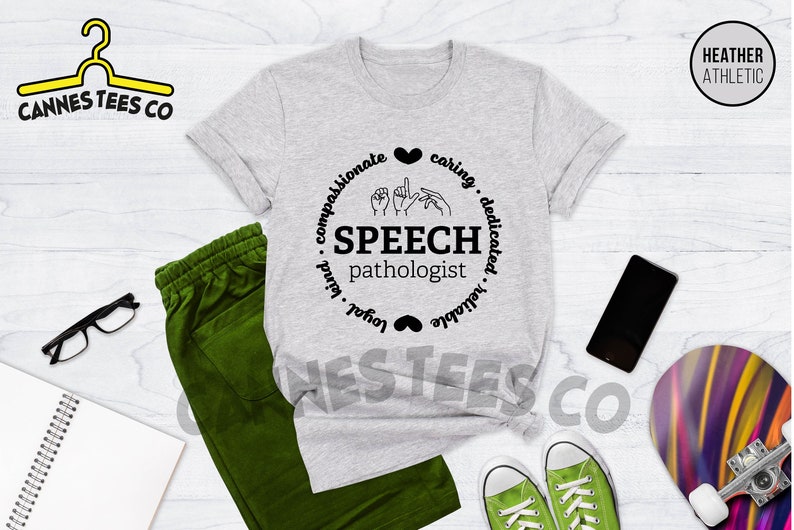 Op de afbeelding: Grijs T-shirt met een zwart cirkelontwerp met de woorden "SPEECH pathologist" en gebarentaal symbolen. De cirkel is omringd door de woorden "compassievol, zorgzaam, toegewijd, betrouwbaar, loyaal, vriendelijk".