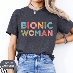 May include: A heather dark grey t-shirt with the text "Bionic Woman" in colorful, distressed lettering.
