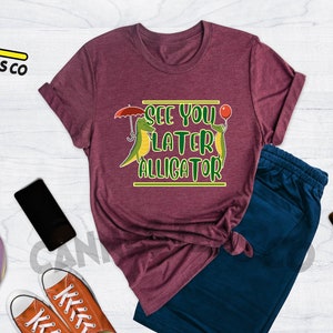 May include: Heather maroon t-shirt with a graphic of two green alligators, one holding an umbrella and the other holding a red balloon. The text "See you later alligator" is printed in yellow on the shirt.