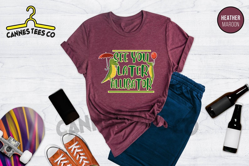 May include: Heather maroon t-shirt with a graphic of two green alligators, one holding an umbrella and the other holding a red balloon. The text "See you later alligator" is printed in yellow on the shirt.