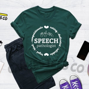 Op de afbeelding: Een groen T-shirt met een wit cirkelvormig ontwerp met de tekst "SPEECH pathologist" met handgebaren voor het woord "speech" en de woorden "compassionate, caring, dedicated, loyal, valuable" rond de cirkel.