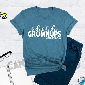 May include: A teal blue t-shirt with the text "i don't do grown ups #pediatrician" in white. The shirt is laid flat on a white wooden surface.