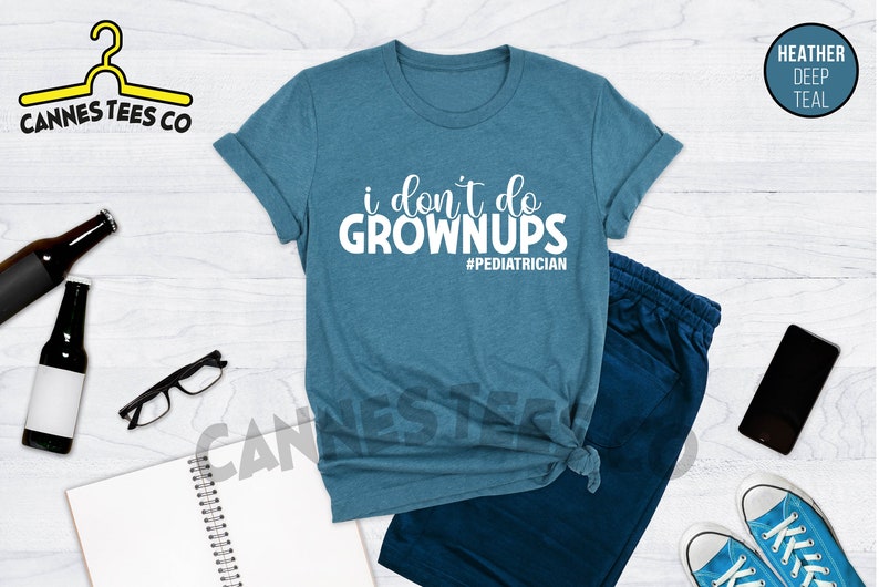 May include: A teal blue t-shirt with the text "i don't do grown ups #pediatrician" in white. The shirt is laid flat on a white wooden surface.